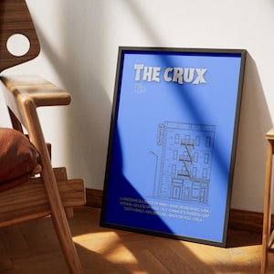 May include: A framed art print with a blue background and the text "THE CRUX" in silver. The print features a line drawing of a building with a fire escape. The print is displayed next to a wooden chair.