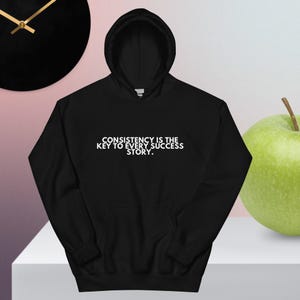 May include: Black hooded sweatshirt with the text "CONSISTENCY IS THE KEY TO EVERY SUCCESS STORY" in white. The sweatshirt has a front pocket and a drawstring hood. A green apple is on the right side of the sweatshirt.