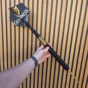 May include: A large dart featuring a black flight with the white Guinness logo and a gold harp emblem. The shaft is black with gold rings, and the barrel has a textured black grip with a gold tip. The dart is held in a hand against a wood wall.