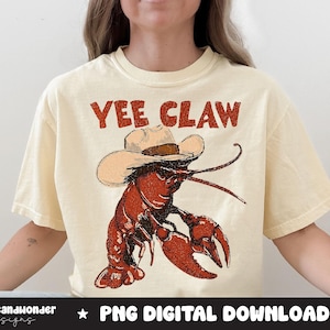 May include: Cream-colored t-shirt featuring a cartoon lobster wearing a cowboy hat. The lobster is red and brown, with the words "Yee Claw" in red above it. The shirt is a digital download.