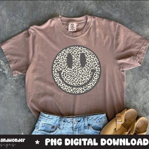 May include: A mauve-colored t-shirt featuring a large smiley face with a leopard print design. The shirt is paired with denim shorts and tan sandals. The text "PNG DIGITAL DOWNLOAD" is visible at the bottom of the image.