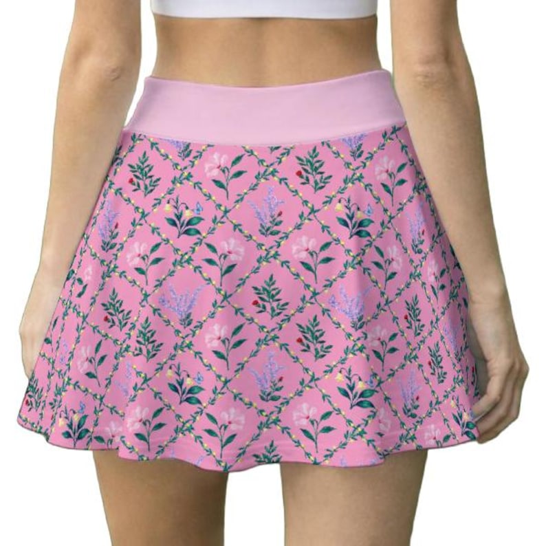 Peut inclure: Une jupe patineuse rose orn&eacute;e d'un motif en losanges r&eacute;p&eacute;t&eacute;, form&eacute; par des vignes vertes. Le design comprend des illustrations de fleurs roses, de feuilles vertes et de petits brins violets dans chaque section, avec une ceinture rose unie.