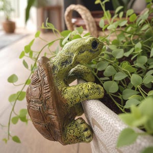 Puede incluir: Una figura decorativa de tortuga en tonos verdes y marrones, agarrada al borde de una maceta. La tortuga tiene un caparazón texturizado y detalles intrincados, añadiendo un toque caprichoso a la disposición de la planta. La figura está hecha de un material duradero.