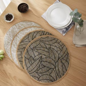 Op de afbeelding: Vier ronde placemats met een bladerenpatroon in beige en zwart liggen op een houten tafel. Twee witte borden, koffiebonen en een kleine vaas met groen staan ook op de tafel.