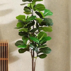 May include: A tall artificial Fiddle Leaf Fig tree in a cream-colored, ribbed planter. The tree has multiple branches with large, vibrant green leaves. The trunk is a dark brown color, and the plant is set against a neutral-toned wall.