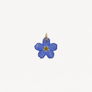 Puede incluir: Un pequeño colgante de flor azul de cinco pétalos con un centro amarillo, que presenta una anilla de metal dorado unida en la parte superior. La flor tiene un acabado liso y brillante, con forma de nomeolvides, sobre un fondo blanco liso.
