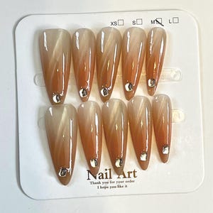 May include: Set of ten almond-shaped false nails with a gradient design, transitioning from a light beige to a warm orange hue. Each nail features a small, silver-colored gem at the base. The card reads "Nail Art".