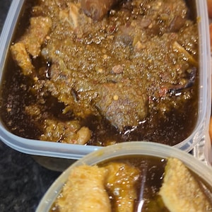 May include: A close-up shot of two clear plastic containers filled with a stew-like dish. The top container holds a dark, thick stew with chunks of meat and a green, speckled sauce. The bottom container has a similar stew with large, golden-brown pieces.