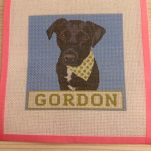 May include: A square needlepoint canvas featuring a black dog wearing a green and white patterned bandana against a light blue background. Below the dog, a rectangular green border contains the name GORDON in white block letters. The canvas has a pink border.