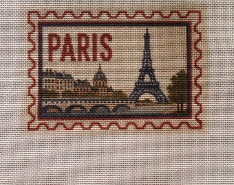 May include: A needlepoint canvas featuring a rectangular postage stamp design with a scalloped red border. The design depicts the Eiffel Tower, a bridge, and city buildings in dark blue and brown tones, with the word PARIS in red text.