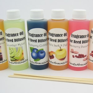 May include: Seven small glass bottles with white lids filled with different colored liquids. Each bottle has a label that reads "Fragrance Oil for Reed Diffusers" with a different scent listed below. The scents are: Hot Cider & Chestnut, Cucumber Melon, Blueberry Cobbler, Apple Jack 'N Pie, Cranberry Peppermint, Candy Cane, and Gingerbread.  The bottles are arranged in a row on a white surface. There are also six wooden sticks in the image.