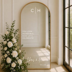 May include: An elegant, arched mirror with a gold frame. The mirror features white text: "YOU LOOK INCREDIBLE, NOW LET'S CELEBRATE!" and the names "Chris & Hannah" with a date. A floral display of white roses and greenery is on the left.