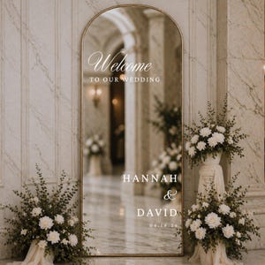 May include: An arched mirror with a gold frame, featuring the text "Welcome TO OUR WEDDING". Below, the names "HANNAH & DAVID" and the date "06.18.26" are displayed. The mirror is surrounded by floral arrangements, creating a wedding theme.