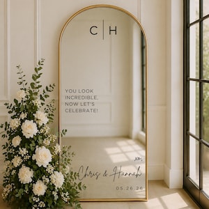 May include: An arched gold mirror with the initials "C | H" at the top. The mirror has the text "YOU LOOK INCREDIBLE, NOW LET'S CELEBRATE!" and the names "Chris & Hannah" with the date "05.26.26" written on it. A floral arrangement is on the left.