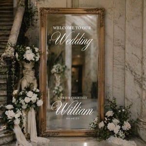 May include: A large, gold-framed mirror with the words "Welcome to Our Wedding" in script. The names "Chris & Courtney William" and the date "04.06.23" are below. The mirror is surrounded by floral arrangements, creating a welcoming wedding display.