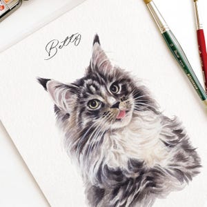 以下が含まれることがあります： グレーと白の毛並みで舌を出したふわふわの猫の水彩画。猫の名前「Betty」は筆記体で書かれています。背景には2本の筆があります。