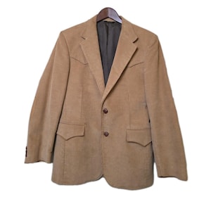 May include: A tan corduroy blazer with a two-button closure, two front pockets, and a notched lapel. The blazer is hanging on a wooden hanger against a white background. The interior lining is a dark green color.
