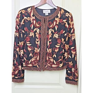 May include: A black long-sleeve jacket featuring an intricate embroidered pattern in shades of red, gold, and tan. The garment has a round neckline, an open front design, and detailed border stitching along the edges. A label reads Laurence Kazar.