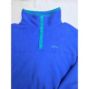 May include: A blue fleece pullover with a high collar and a teal accent on the collar and button placket. The pullover has a small embroidered logo on the chest. The fleece is soft and appears to be a comfortable garment.