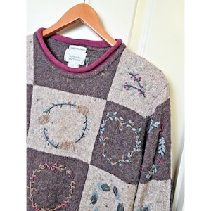 Könnte beinhalten: Ein braun-beiger karierter Pullover mit gestickten Blumenmustern. Der Pullover hat einen bordeauxfarbenen Ausschnitt und hängt an einem Holzbügel. Die Marke des Pullovers ist Christopher & Banks.