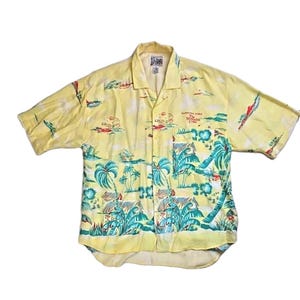 May include: Yellow short-sleeve button-down shirt with a tropical print featuring palm trees, huts, and sailboats. The shirt has a camp collar and a pocket on the chest. Text on the shirt reads "Harrison Ford Mosquito Coast".