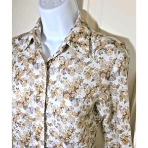 Puede incluir: Una camisa abotonada con estampado floral. La camisa presenta un fondo blanco con un patrón repetido de pequeñas flores amarillas, marrones y moradas. El cuello y los puños son visibles, y la tela parece ligera.