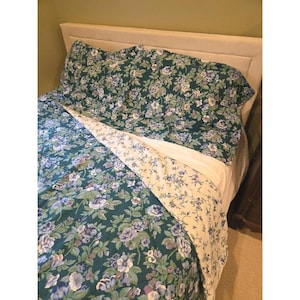 May include: A bed covered with a floral patterned comforter and pillowcases. The comforter features a dark teal background with blue, purple, and white flowers. The underside of the comforter is a cream color with a smaller floral pattern.