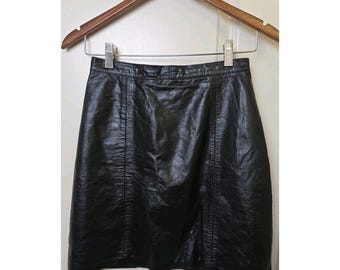 Può includere: Una gonna di pelle nera appesa a una gruccia di legno. La gonna è un classico modello a trapezio con cuciture verticali. La pelle ha una leggera lucentezza. Lo sfondo è una porta bianca.