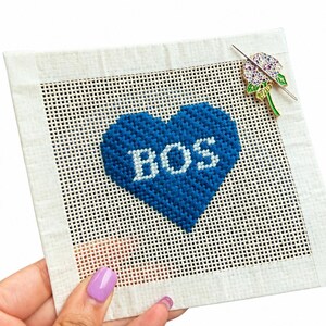 Puede incluir: Un lienzo de punto de aguja cuadrado con un diseño de corazón azul y las letras "BOS" en blanco. Un alfiler decorativo con una flor morada y hojas verdes está adjunto. El lienzo es sostenido por una mano con esmalte de uñas morado.