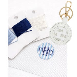 Op de afbeelding: Witte leren AirTag-houder met goudkleurige hardware en een sleutelring. De houder is rond met een gestikte rand. Ook afgebeeld zijn spoelen met blauw en wit borduurgaren en een kruissteekontwerp met de initialen "MHD".