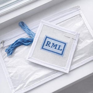 May include: A white needlepoint canvas with the letters "RML" stitched in blue, a needle, and blue thread. The canvas is inside a clear zippered pouch. The items are on a white surface near a window.