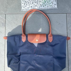 May include: A navy blue tote bag with brown leather handles and a flap closure. The bag has a rectangular shape and a gold-colored button. A white bag with the word "CHAMP" repeated is visible in the background.