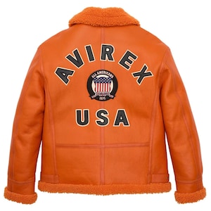 ack view of a handmade men's vibrant orange leather B3 bomber jacket featuring large black and white AVIREX USA varsity lettering and an American flag patch, with plush orange shearling fur trimming on the collar and hem.