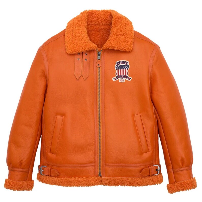 Front view of an orange aviator shearling jacket showing a heavy-duty zipper closure, thick orange fur collar, an American flag shield patch on the left chest, dual throat straps, and adjustable waist buckles.