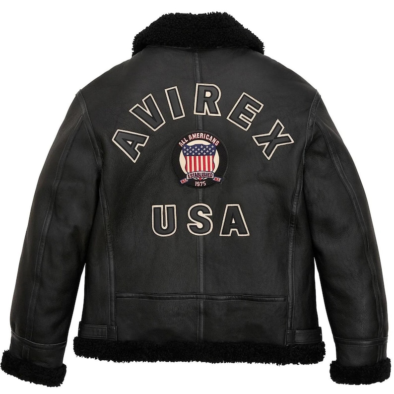 May include: Black leather jacket with a shearling collar and trim. The back features the word "AVIREX" in white lettering, along with a circular patch displaying the American flag and the text "ALL AMERICANS ESTABLISHED 1975 USA".