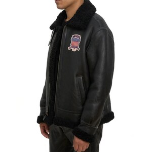May include: Black leather jacket with a black shearling collar and trim. The jacket features a zipper closure and a patch on the chest with the text "AVIREX" and an American flag design. The jacket has a classic aviator style.