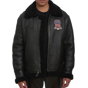 May include: Black leather jacket with a shearling collar and lining. The jacket features a zipper closure, a patch with an American flag design, and the word "AVIREX". The jacket has a classic aviator style.