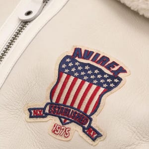 May include: Close-up of a white leather jacket with a prominent Avirex patch. The patch features a shield design with the American flag, the text "AVIREX", and "ESTABLISHED 1975 NY". A white zipper and a leather pull are visible.