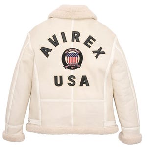 May include: Cream-colored shearling jacket with a sheepskin collar and cuffs. The back features the word "AVIREX" above a circular emblem with the words "All Americans" and "USA" below. The emblem includes a red, white, and blue design.