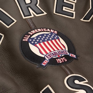 Close-up of the upper back on a brown leather jacket highlighting the detailed stitching on the All Americans 1975 shield patch and varsity letters.