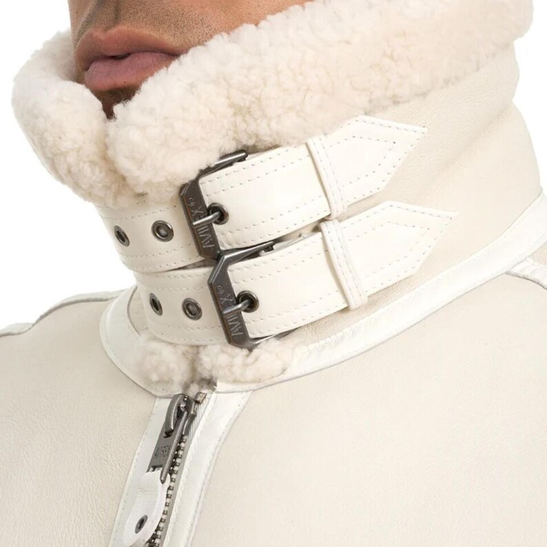 May include: Close-up of a cream-colored jacket with a shearling collar. The jacket features white leather straps with silver buckles and a zipper closure. The collar is lined with soft, white shearling.