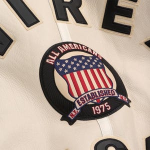 May include: Close-up of a cream-colored jacket with black and white lettering. The jacket features a circular patch with an American flag design, the words "ALL AMERICANS" and "ESTABLISHED 1975".