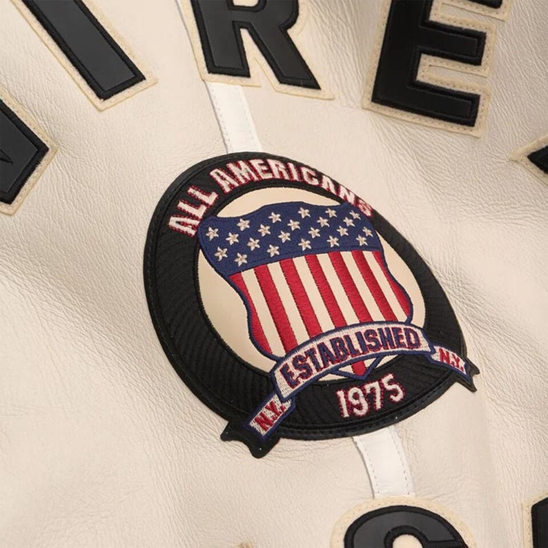 May include: Close-up of a cream-colored jacket with black and white lettering. The jacket features a circular patch with an American flag design, the words "ALL AMERICANS" and "ESTABLISHED 1975".