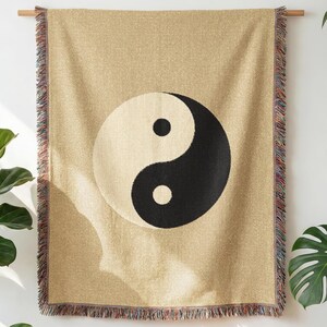 May include: A beige throw blanket featuring a large black and white yin yang symbol. The blanket has a fringed edge with multiple colours. It is suspended from a wooden rod.