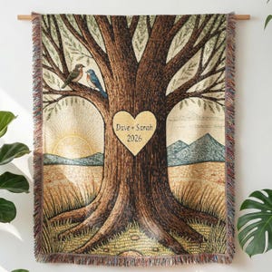 Op de afbeelding: Geweven sprei met een boomontwerp, vogels en een hart met "Dave + Sarah" en 2026. De deken heeft een franjerand en toont een zonsopgang en bergen. De afmetingen zijn in centimeters.