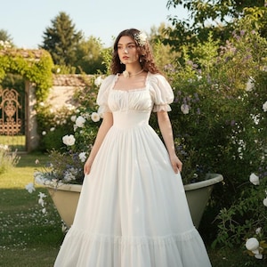 May include: A white, floor-length dress with a square neckline and short, puffed sleeves. The dress features a fitted bodice and a flowing skirt with a ruffled hem. The background includes a garden setting with white roses and a decorative archway.