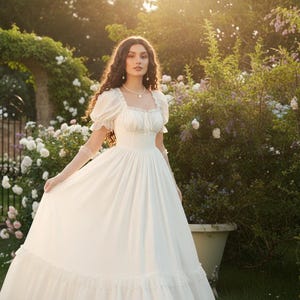 May include: A woman in a white, vintage-style dress with a square neckline and puff sleeves stands in a garden. The dress has a full skirt and is accessorized with a necklace and gloves. The background features a stone archway, roses, and lush greenery.