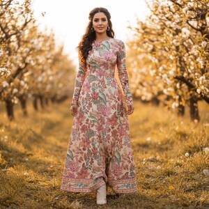 May include: A long-sleeved, floral-print dress in shades of pink, green, and cream. The dress features a round neckline and a flowing skirt. The person is standing in a field of tall grass with trees in the background.