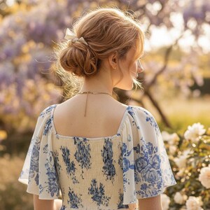 May include: A woman's dress with a square neckline and short sleeves, featuring a blue floral pattern on a cream background. Her hair is styled in a bun with a white bow. The background includes white roses and purple flowers.