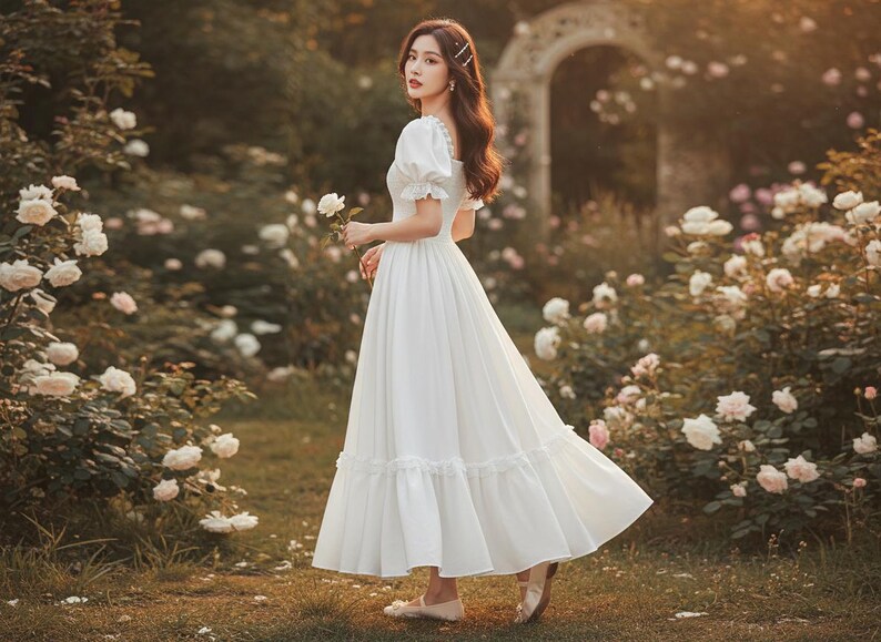 May include: A long, white dress with short, puffed sleeves and a flowing skirt. The dress features a ruffled hem and is worn in a garden setting with white roses. The dress is a classic, romantic style.